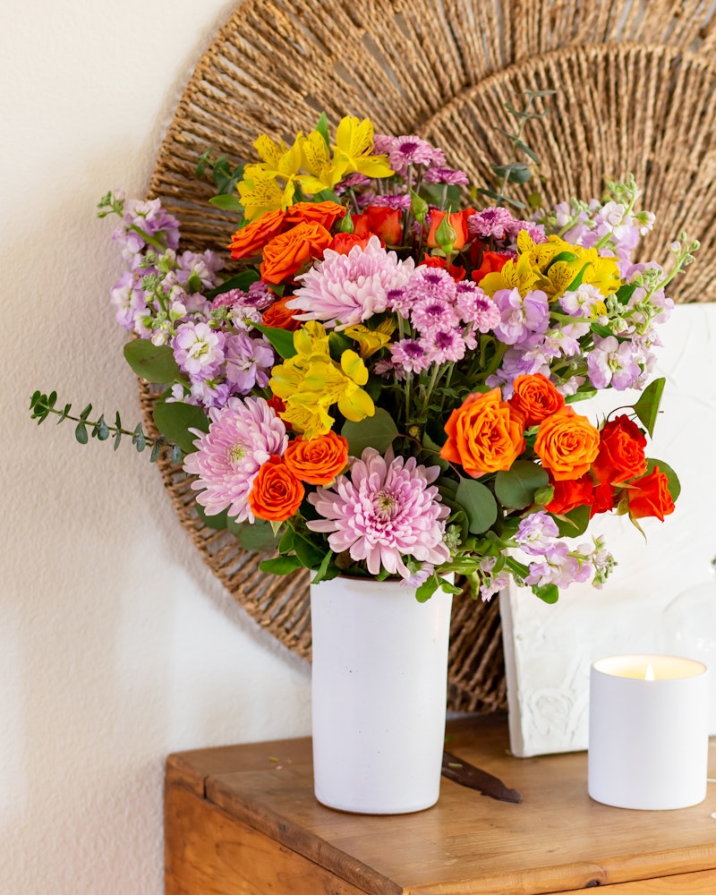 Vibrant bouquet of flowers featuring orange roses, pink chrysanthemums, and yellow blossoms in a white vase on a wooden surface, with a candle and woven decor in the background.