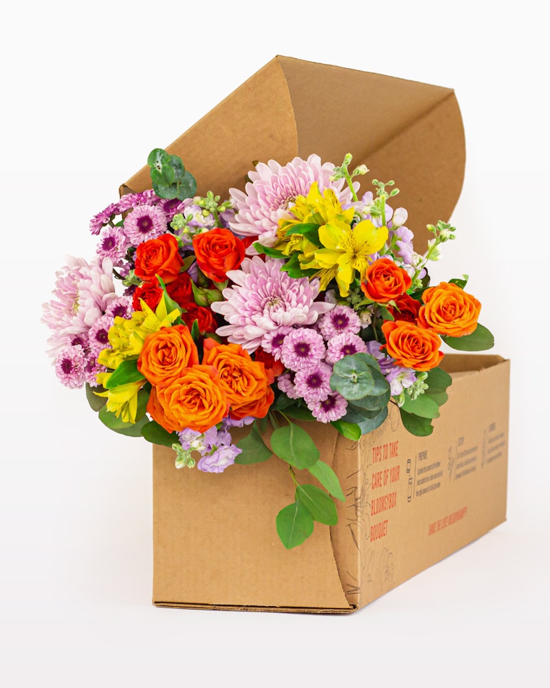 Vibrant bouquet of fresh flowers with roses, daisies, and other mixed blooms packaged neatly in an open cardboard box on a white background.