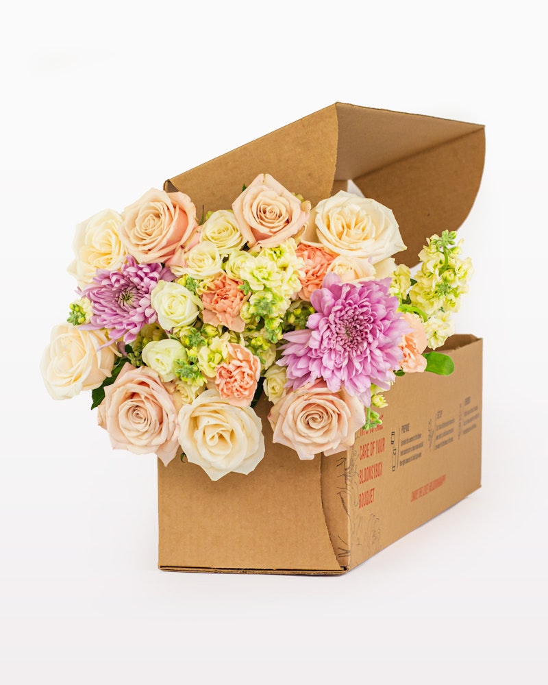 A beautifully arranged bouquet of pastel roses, dahlias, and assorted flowers bursting from an open cardboard gift box on a white background.