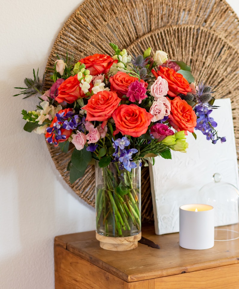Vibrant bouquet of roses and assorted flowers in a glass vase on a wooden table, complemented by a lit candle and decorative wicker plate on a white wall background.