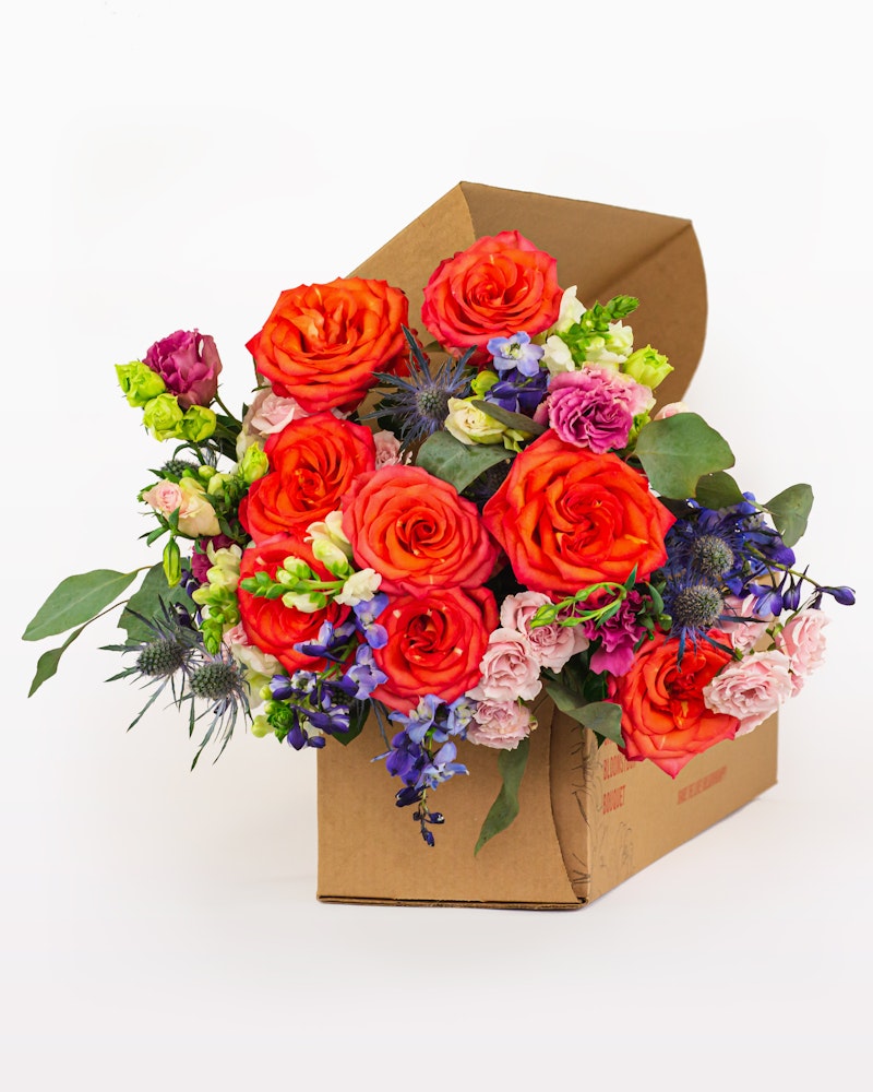 Vibrant bouquet of flowers including bright orange roses, purple blooms, and lush greenery, all packaged neatly in a brown cardboard box against a white background.