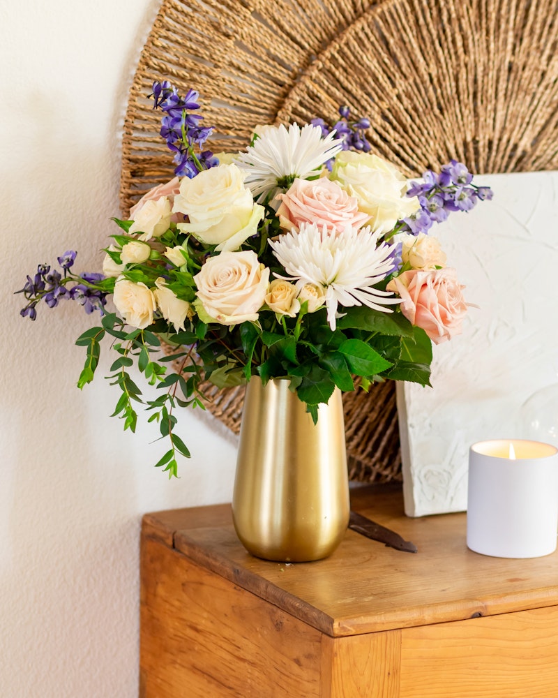 Beautiful bouquet of mixed flowers including roses and chrysanthemums in a gold vase beside a lit candle, set on a wooden table against a white wall with wicker decor.