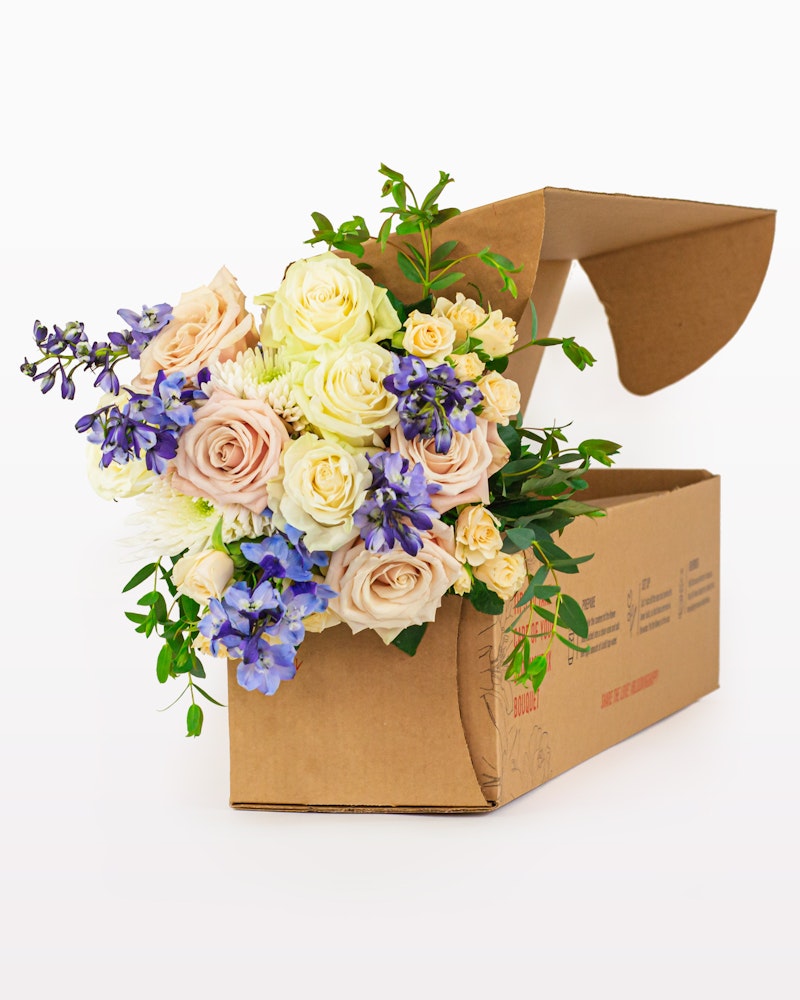 A vibrant bouquet of fresh flowers featuring roses and lavender spilling out of an open cardboard delivery box on a white background, indicating a flower delivery service.