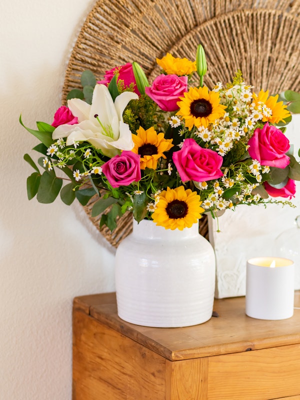Vibrant bouquet of flowers featuring pink roses, sunflowers, and white lilies in a white vase, with a woven fan in the background on a wooden surface next to a lit candle.