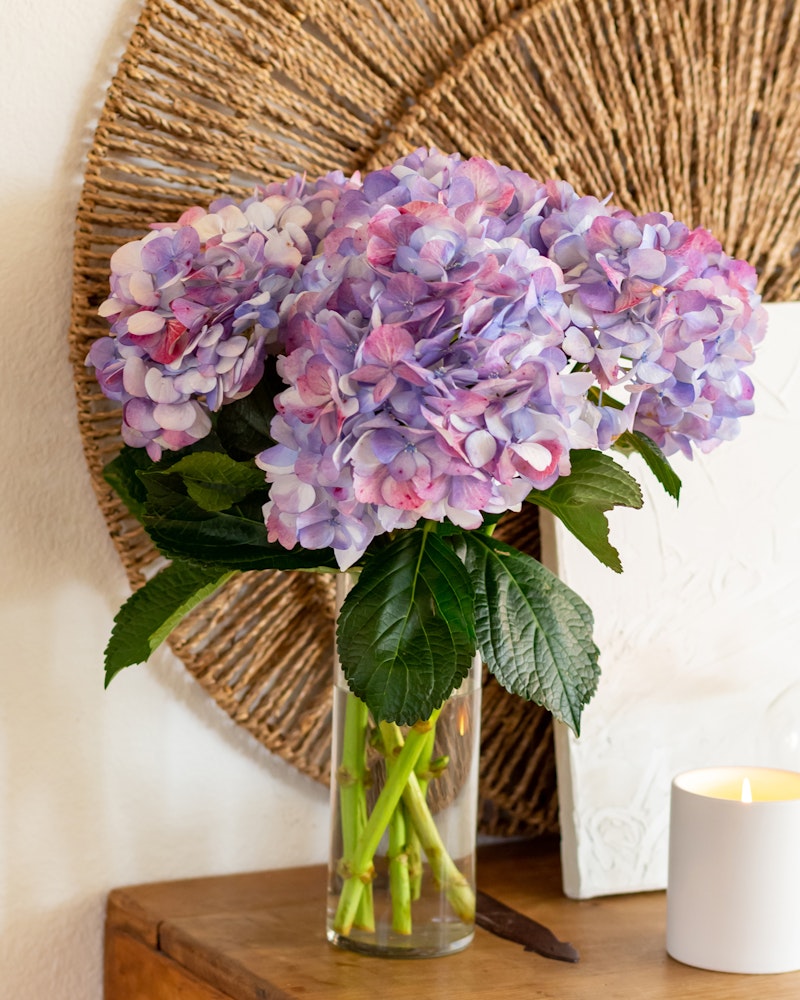 A vibrant bouquet of pink and purple hydrangeas arranged in a clear glass vase on a wooden side table, with a lit candle and a woven wall decoration in the background.
