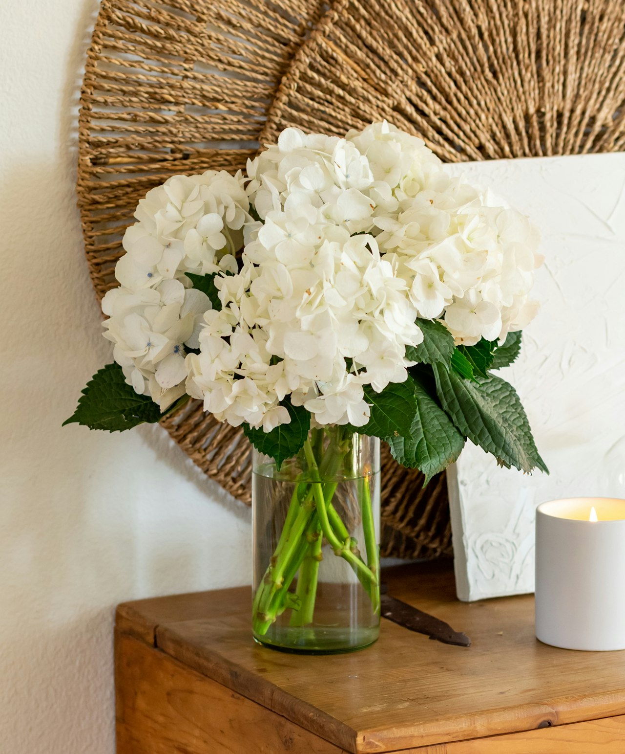 A beautiful arrangement of white hydrangeas in a glass vase, complemented by lush green leaves.