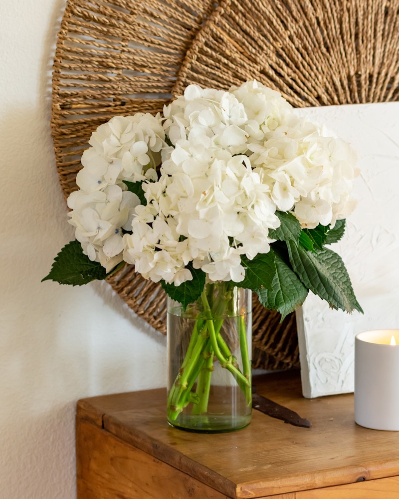 Elegant white hydrangea bouquet in a clear glass vase on a wooden table, with a woven circular wall decor in the background and a lit candle to the side.