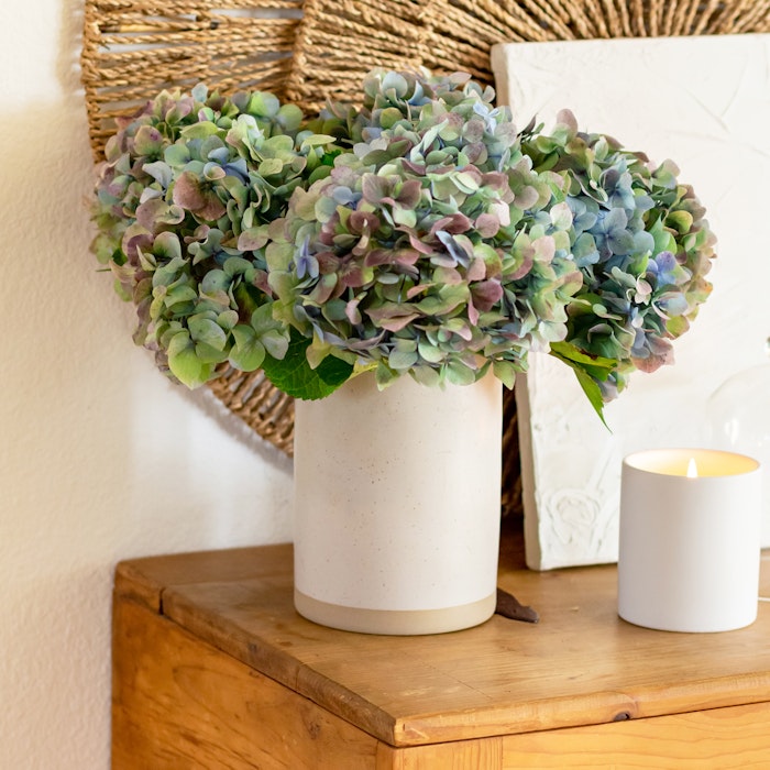 Large bouquet of blue and green hydrangeas in a white speckled vase on a wooden table, with a lit candle and woven decorations in the background.