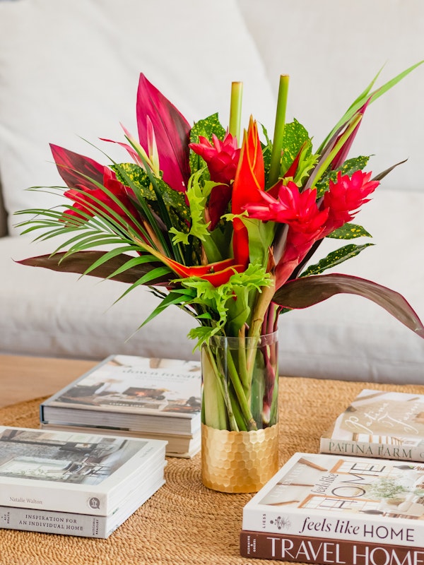 A vibrant bouquet of red flowers and green leaves in a glass vase on a wooden table, with home and travel-themed coffee table books in the background.