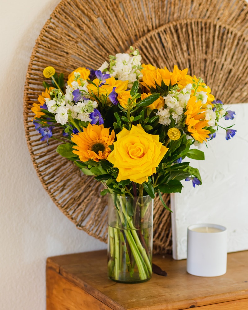 A vibrant bouquet of yellow roses and sunflowers paired with purple flowers, displayed in a clear vase on a wooden table against a white wall and decorative wicker art.