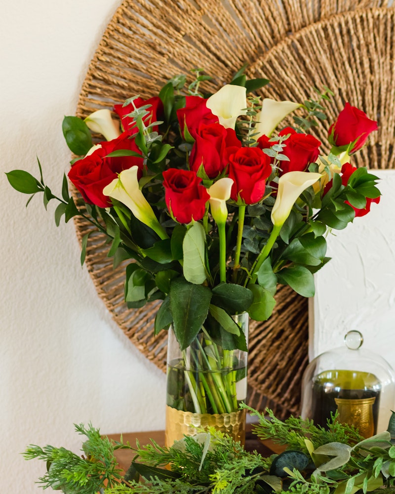 Beautiful bouquet of red roses and white calla lilies in a clear glass vase on a table, with a woven circular wall decoration in the background.