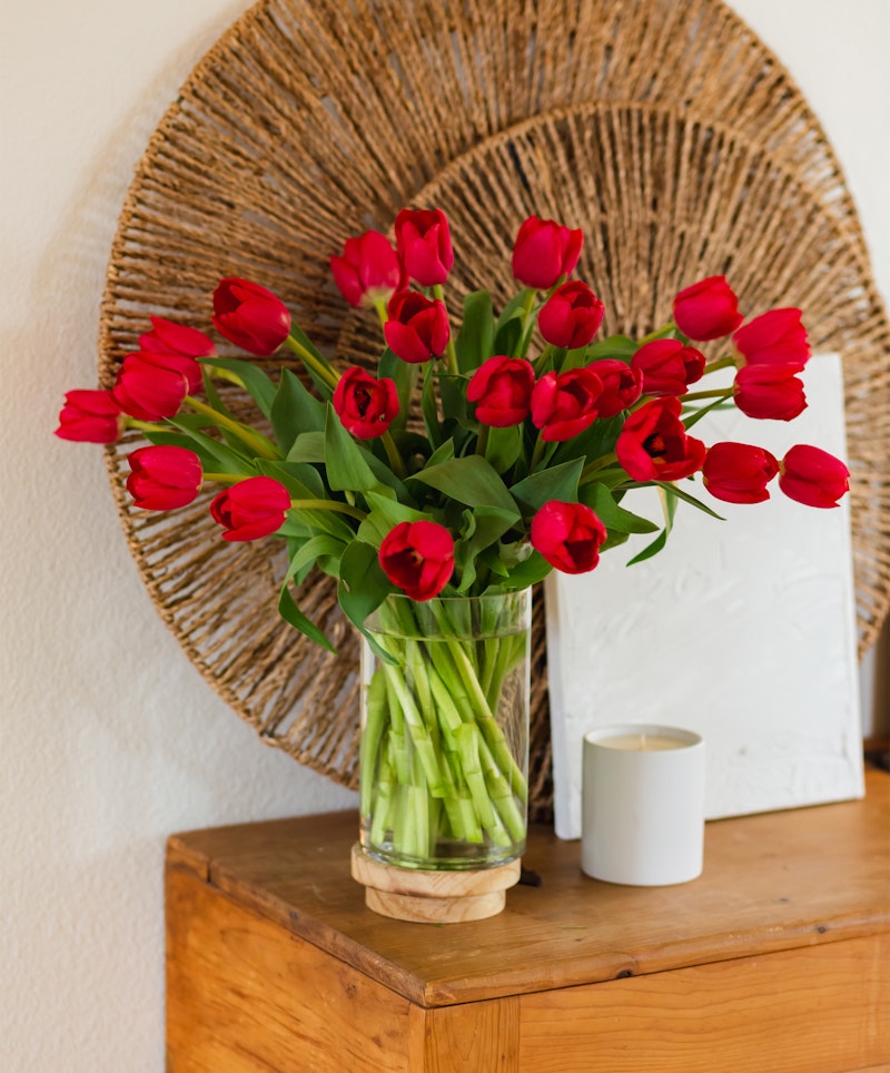 Bouquet of vibrant red tulips in a clear glass vase on a wooden surface with a woven circular wall decor and a white candle beside it, showcasing a cozy home interior.