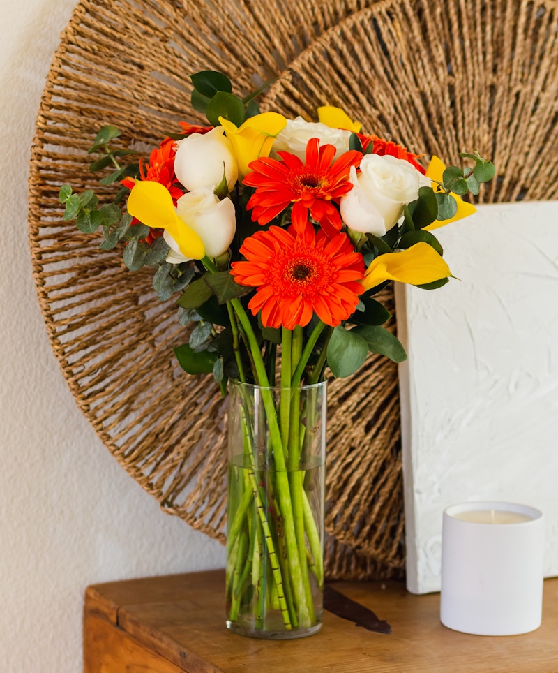 Vibrant bouquet of flowers with red gerberas, white roses, and yellow lilies in a clear vase on a wooden table against a round woven backdrop.