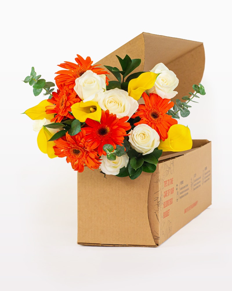 Brightly colored bouquet of orange gerberas, white roses, and yellow lilies spilling from an open cardboard box against a white background, showcasing a flower delivery concept.