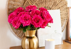 Vibrant pink roses arranged in a golden vase on a table, with a decorative wicker plate and a lit white candle in a minimalist interior setting.