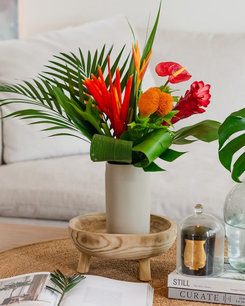 Vibrant tropical flower arrangement in a white vase on a wooden pedestal table, with a cozy beige sofa, books, and a glass hourglass in the background.