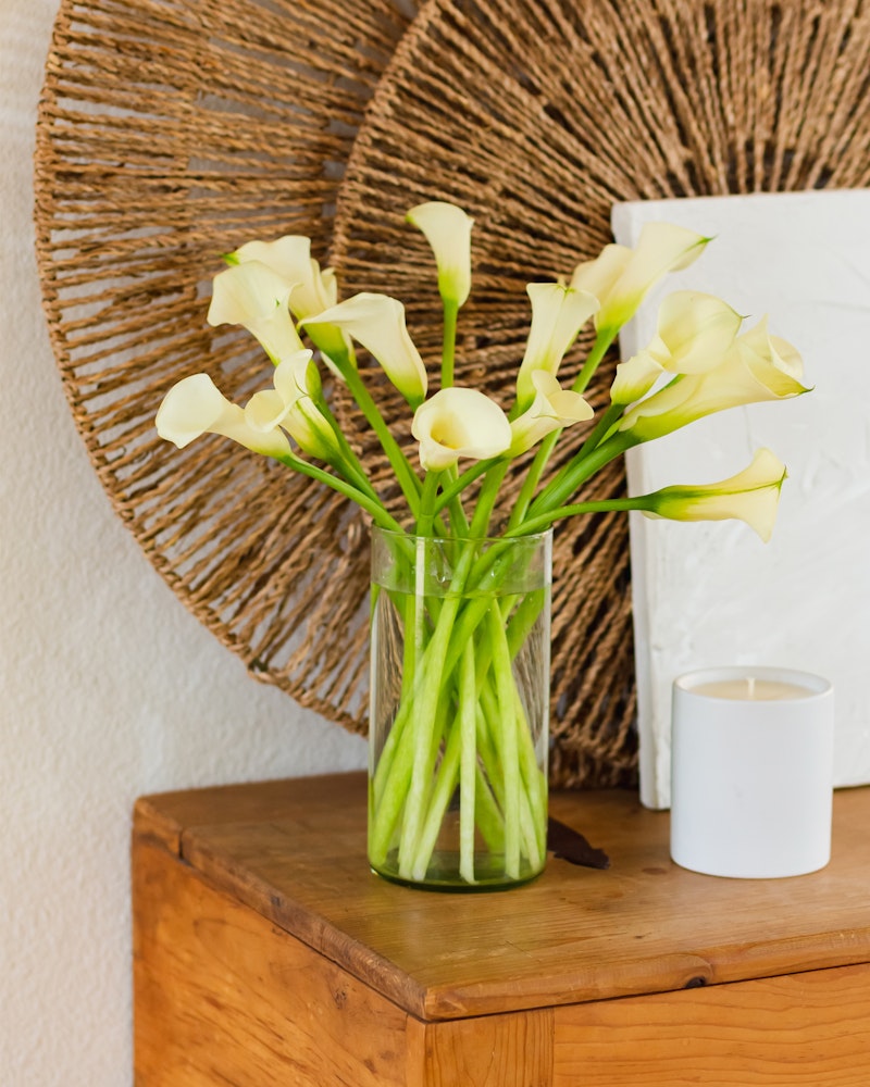 A clear glass vase filled with elegant yellow calla lilies on a wooden table, with a woven circular wall decor and a white candle beside it, composing a tranquil home setting.