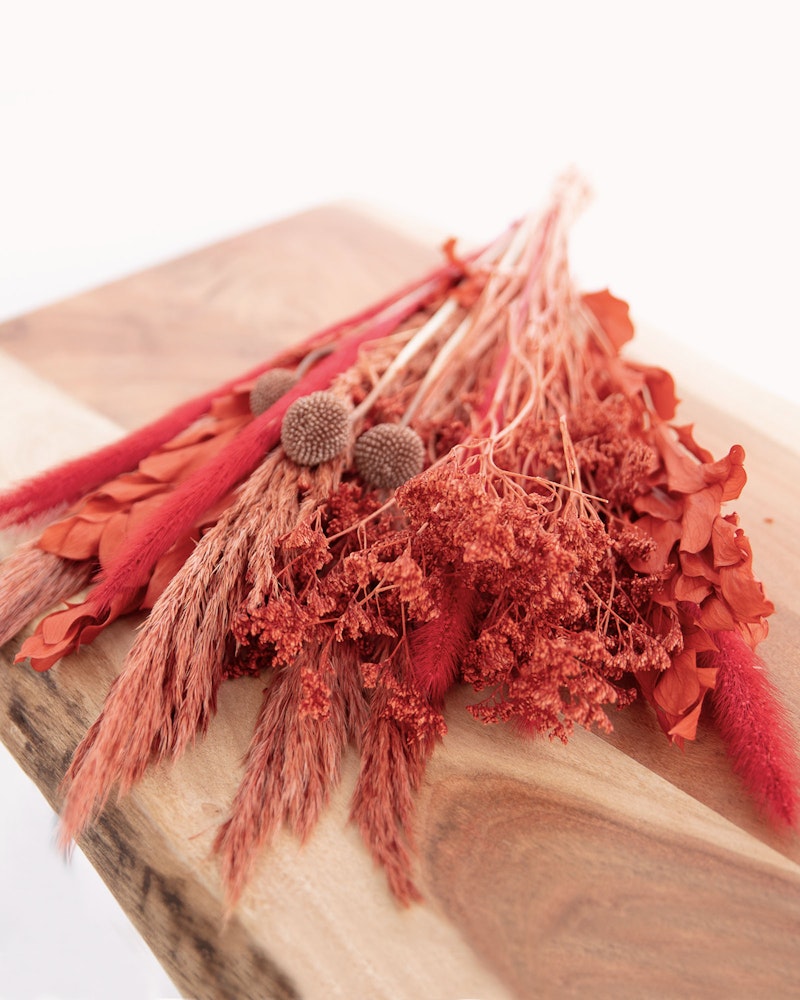 A vibrant bouquet of dried flowers in shades of red and pink, with feathery textures and spherical accents, arranged on a wooden cutting board against a white background.