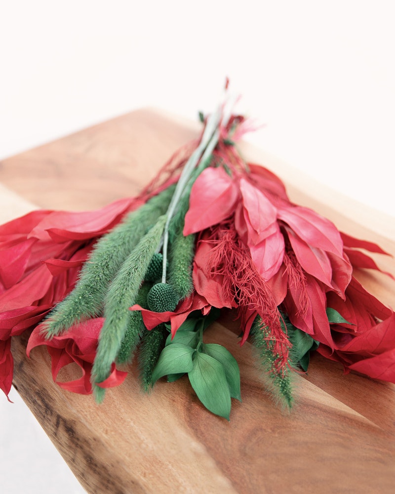 Vibrant bouquet of red flowers and greenery, featuring unique textures, laid on a smooth wooden surface with a neutral background enhancing colors.