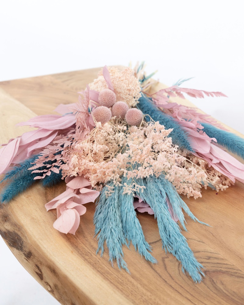 A vibrant bouquet of dried flowers in pastel pinks, corals, and teal blue hues, artistically arranged on a smooth wooden surface against a white background.