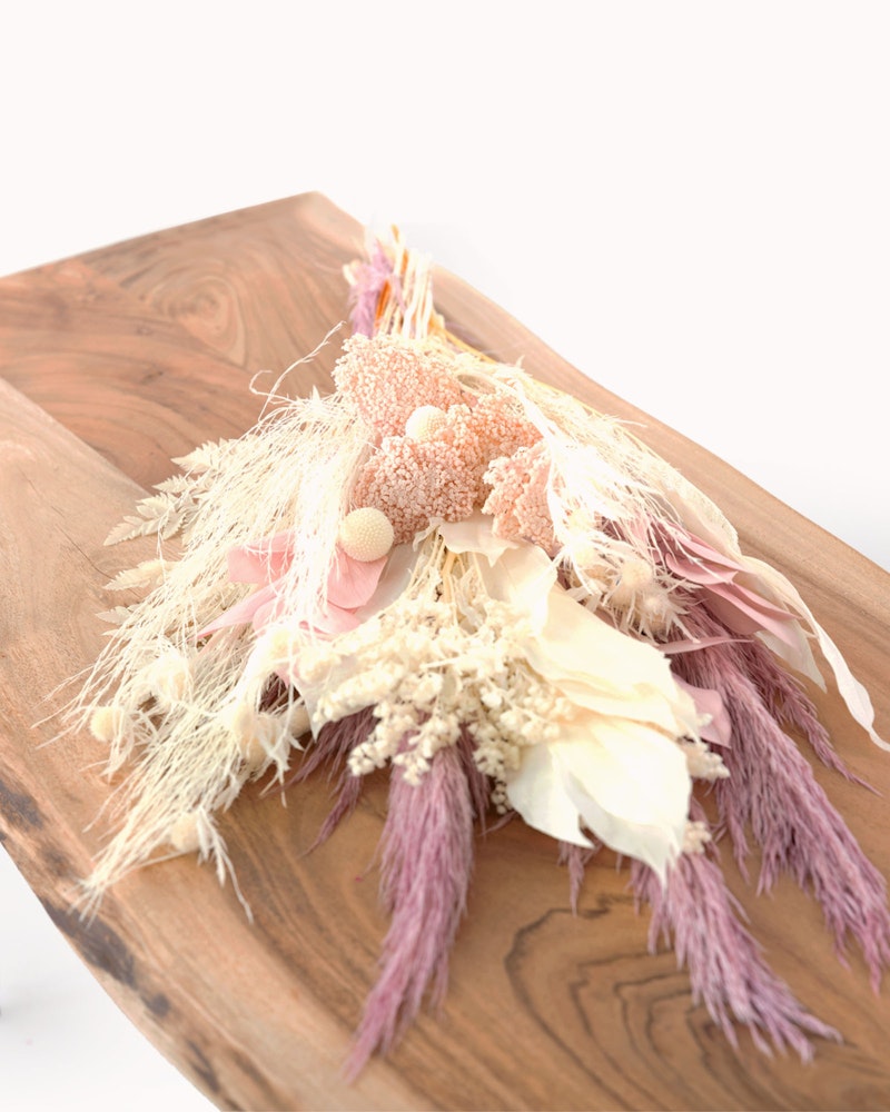 Elegant dried flower arrangement in blush and neutral tones, displayed on a smooth wooden slab with a curved edge, showcasing a mix of textures and organic shapes.