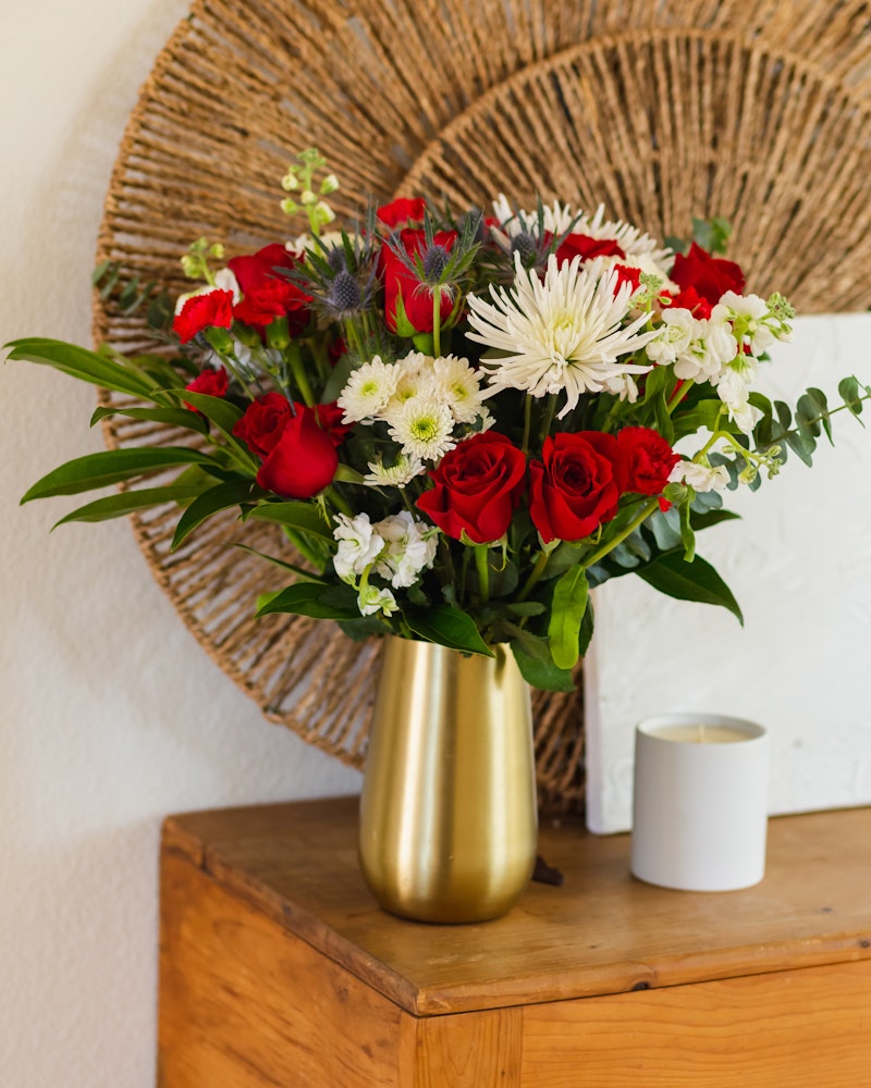Vibrant bouquet of red roses and white chrysanthemums in a golden vase on a wooden table beside a lit candle, with a woven wall decoration in the background.
