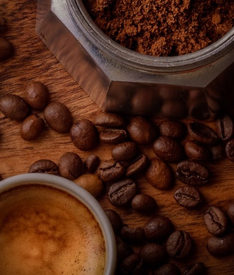 Aromatic espresso in a white cup with scattered coffee beans and a jar of ground coffee on a rustic wooden table, invoking a cozy, warm atmosphere.