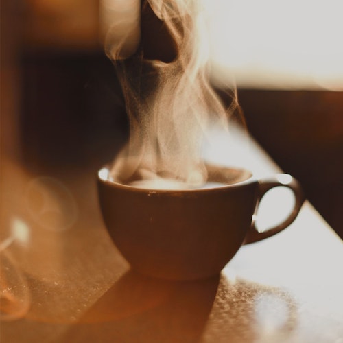 Close-up of a steaming cup of coffee on a wooden surface with warm, natural lighting highlighting the vapor trails rising from the beverage.