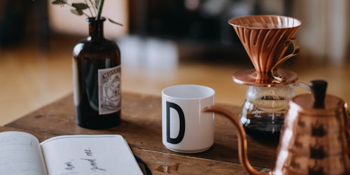Inviting coffee setup with a pour-over brewer, a cup with letter D, a notebook, and a vase on a wooden table, evoking a cozy, creative morning atmosphere.