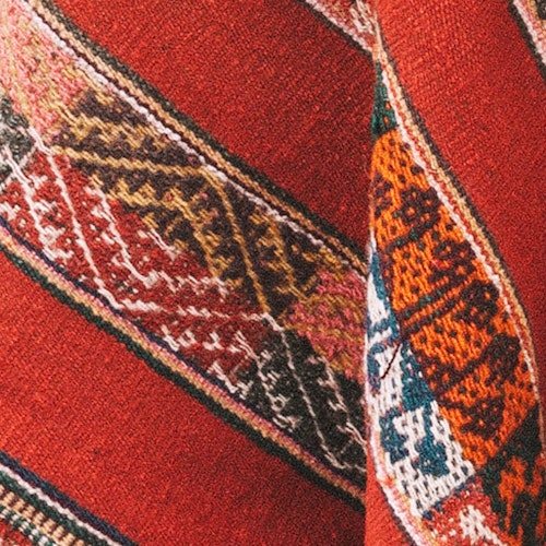 Close-up of a traditional patterned textile with vibrant reds, detailed geometric shapes, and hints of orange and white threading.