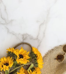 Bright yellow sunflowers in a wicker basket next to stylish sunglasses perched on a straw hat, all arranged on a sleek marble surface with copy space.