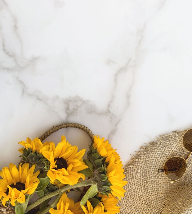 Bright sunflowers arranged on a marble surface next to stylish sunglasses and a straw hat.