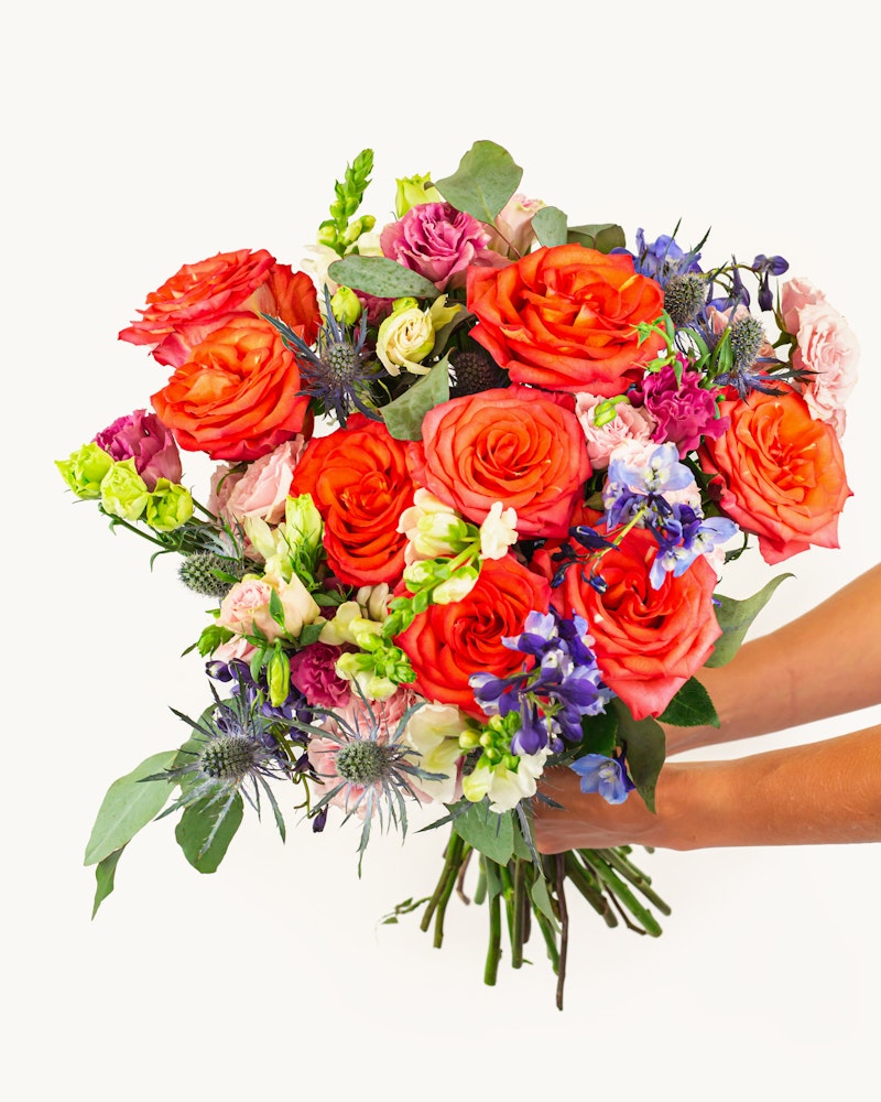 Vibrant bouquet of flowers with red oranges, and purple hues, including roses, snapdragons, and lilies, held up against a white background.