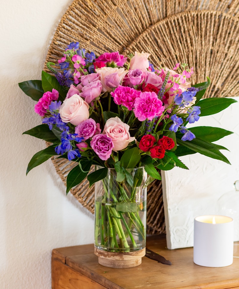 Vibrant bouquet of mixed flowers, including roses and delphiniums in a glass vase, accentuated by green foliage on a wooden table with a candle and wall decor.