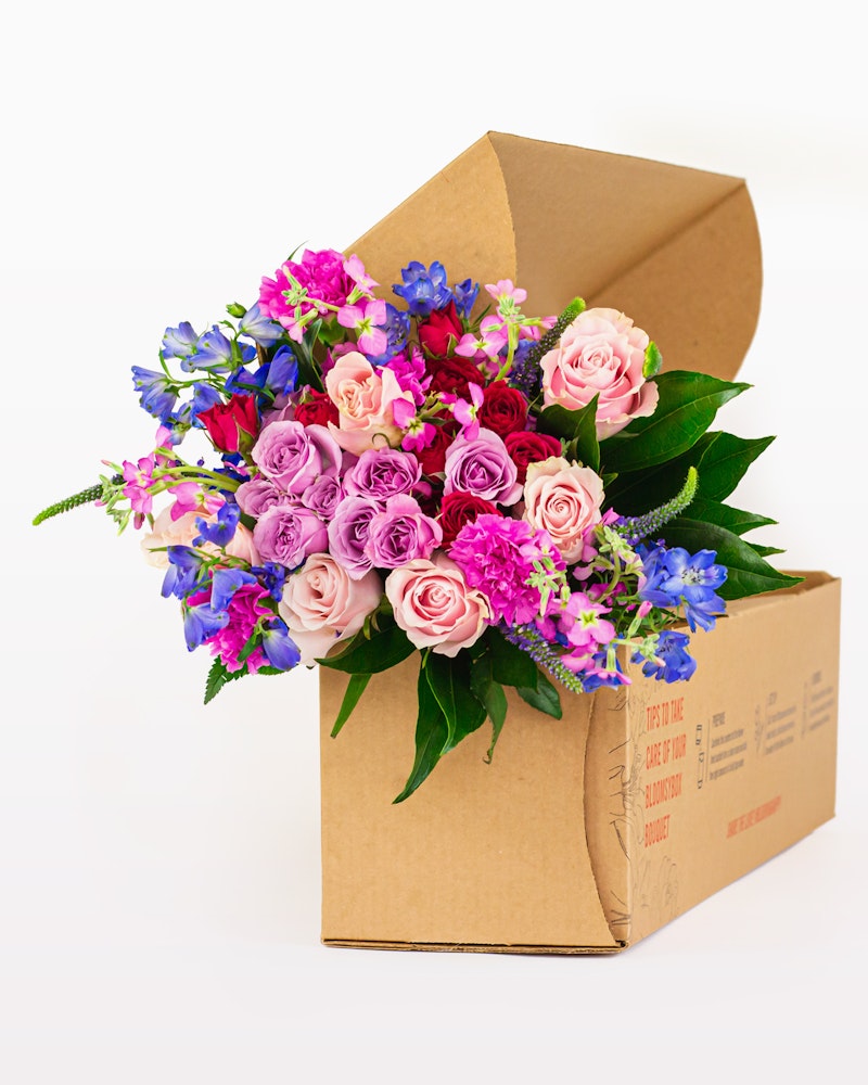 Vibrant bouquet of pink roses, blue flowers, and greenery arranged in a recyclable brown cardboard box on a white background, ideal for eco-friendly gifting.