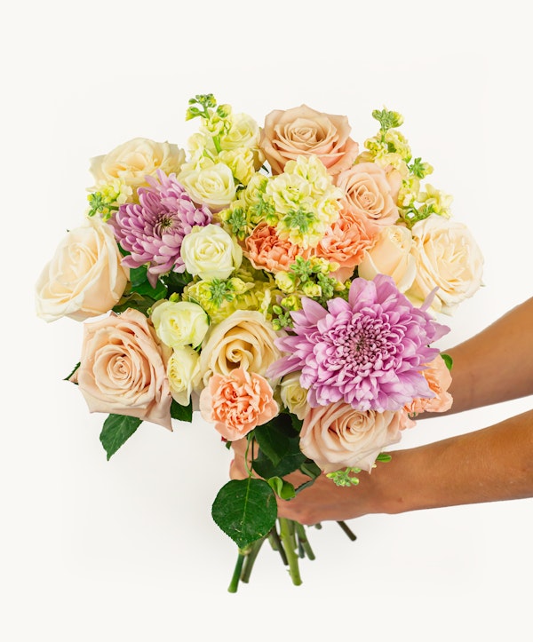 A vibrant bouquet of flowers in shades of pink, peach, and cream, held by a person against a white background, featuring roses, lilies, and other mixed blooms.