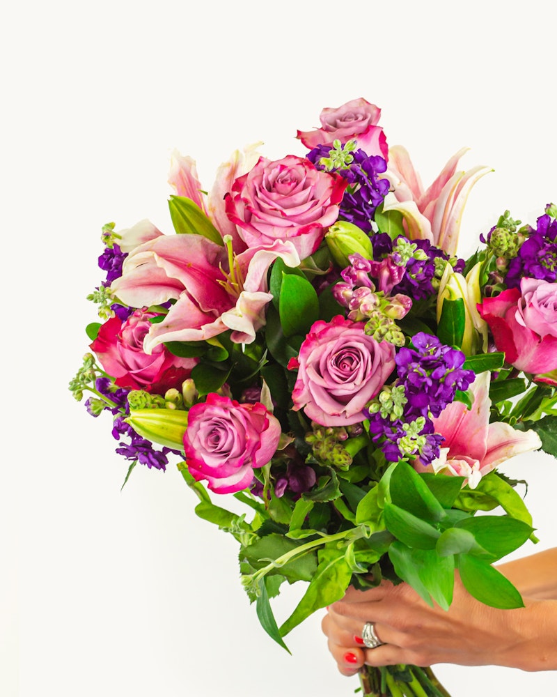 Vibrant bouquet of flowers including pink roses, purple accents, and green foliage held by a person's hand against a white background, showcasing a fresh floral arrangement.