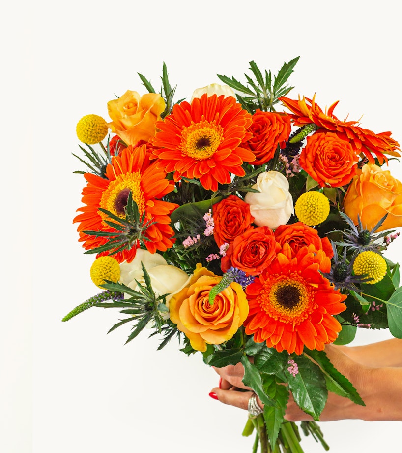 Vibrant bouquet of flowers including orange gerberas, yellow roses, and various greenery held by a person against a white background, focused and brightly colored.