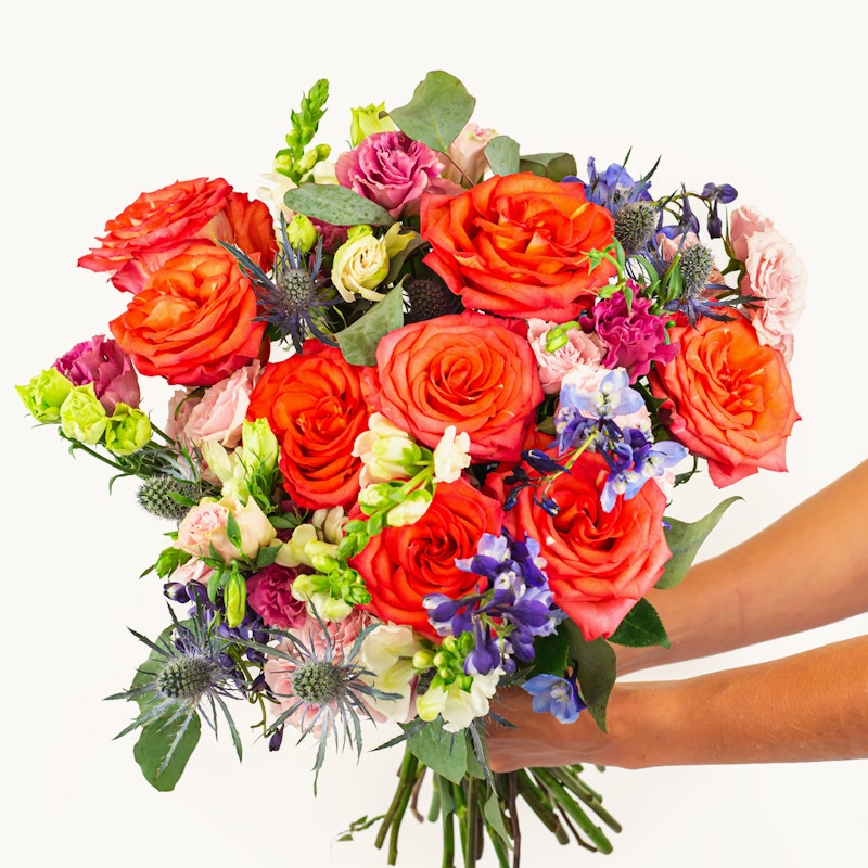 Vibrant bouquet of fresh flowers including orange roses, purple accents, and green foliage held by a person against a white background, symbolizing a gift or celebration.
