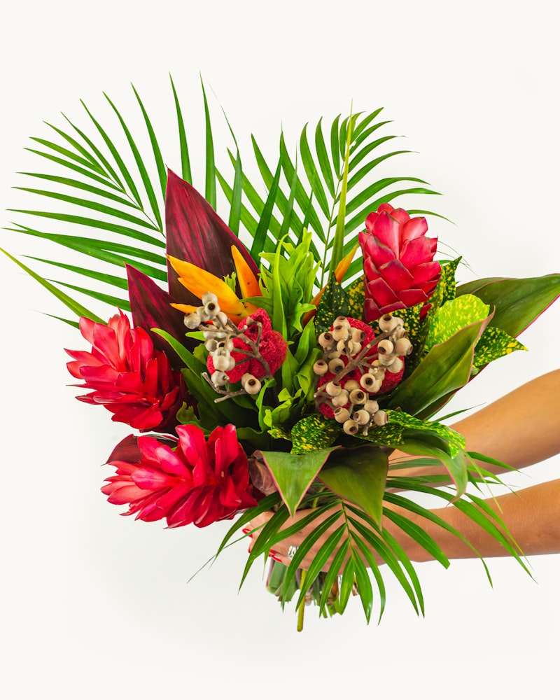 Vibrant tropical bouquet featuring red ginger flowers, lush green palm leaves, exotic foliage, and blooming orchids held against a white background.