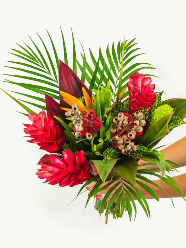 Vibrant tropical bouquet featuring red ginger flowers, lush green palm leaves, exotic foliage, and blooming orchids held against a white background.