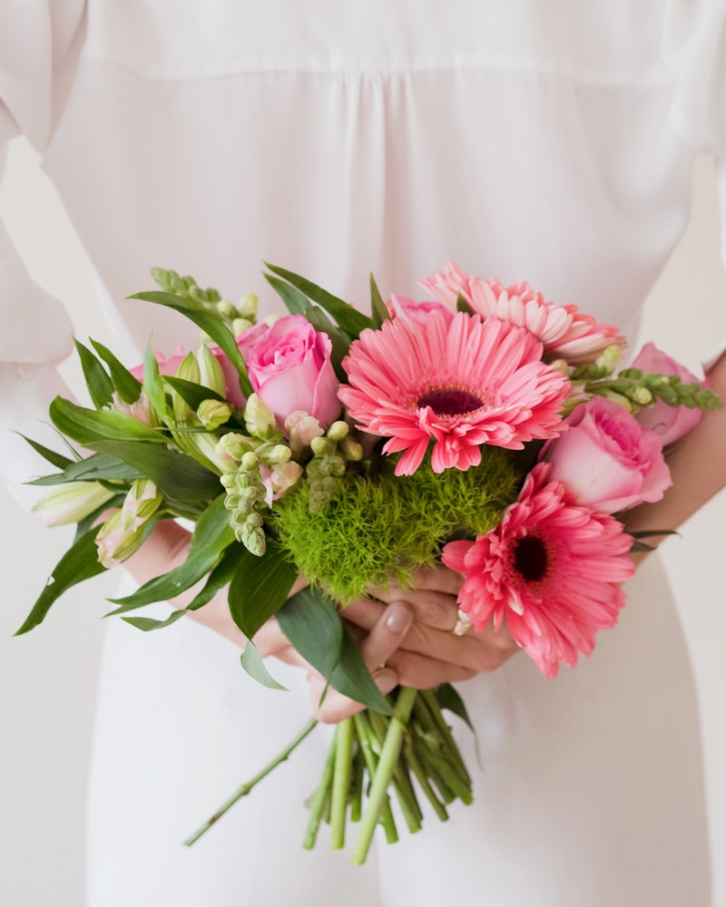 Person in a white dress holding a fresh bouquet of pink roses, gerbera daisies, and green foliage against a plain background, signifying a celebratory or ceremonial event.