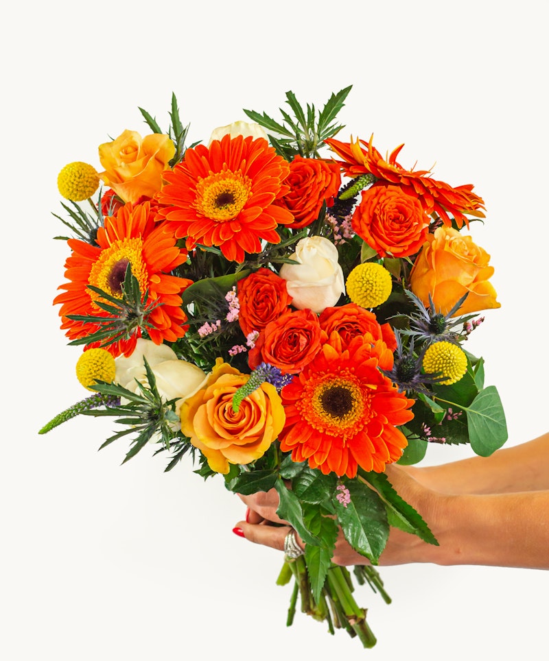 A vibrant bouquet with orange gerberas, yellow roses, and a mix of greenery and filler flowers, cradled in a person's hands against a white background.