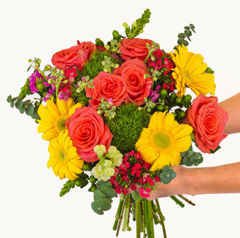 Vibrant bouquet of flowers featuring orange roses, yellow daisies, and various greenery held by a person against a white background, symbolizing freshness and natural beauty.