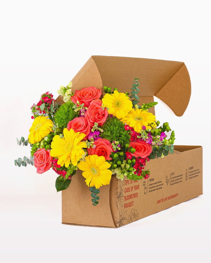 Vivid assortment of fresh flowers, including yellow blooms and red roses, arranged beautifully in a brown cardboard box ready for delivery against a white background.