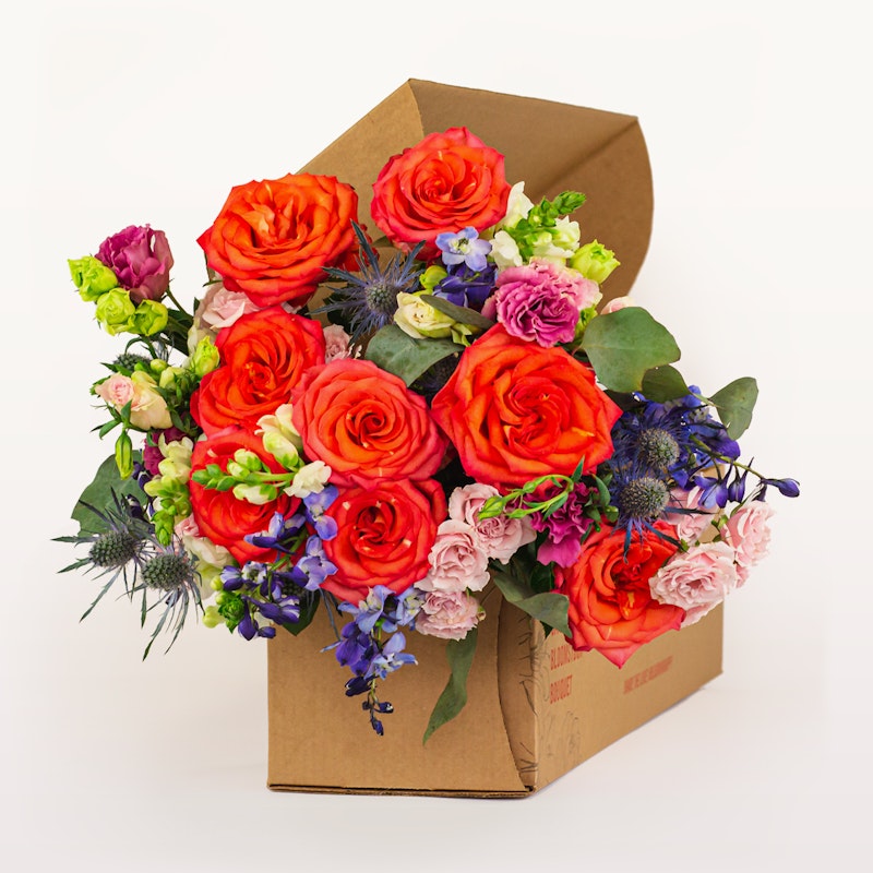 Vibrant bouquet of flowers featuring orange roses, pink blooms, and purple accents, artistically arranged in an open brown cardboard box set against a white background.