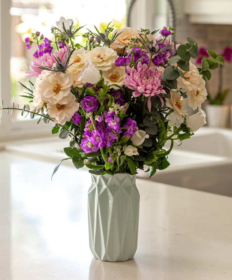 A vibrant bouquet of pink, purple, and white flowers arranged in a geometric pale green vase, sitting on a white countertop against a kitchen window backdrop.