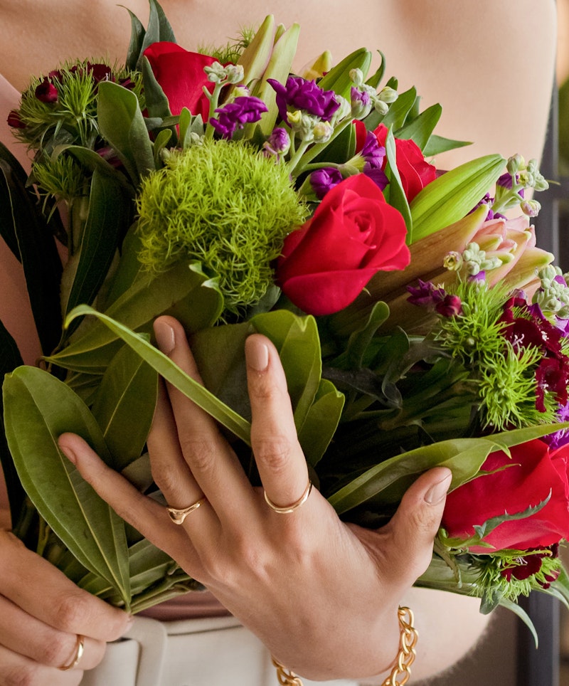 Close-up of a woman's hands cradling a vibrant bouquet of red roses and greenery with a glimpse of her beige dress in the background.