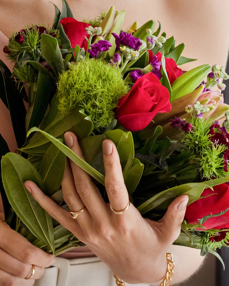 Close-up of a woman's hands cradling a vibrant bouquet of red roses and greenery with a glimpse of her beige dress in the background.