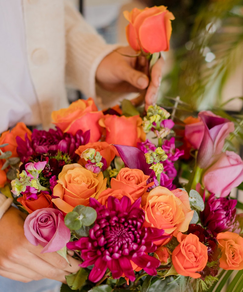 Person in a white sweater arranging a vibrant bouquet of flowers, including orange roses, purple blooms, and lush greenery.